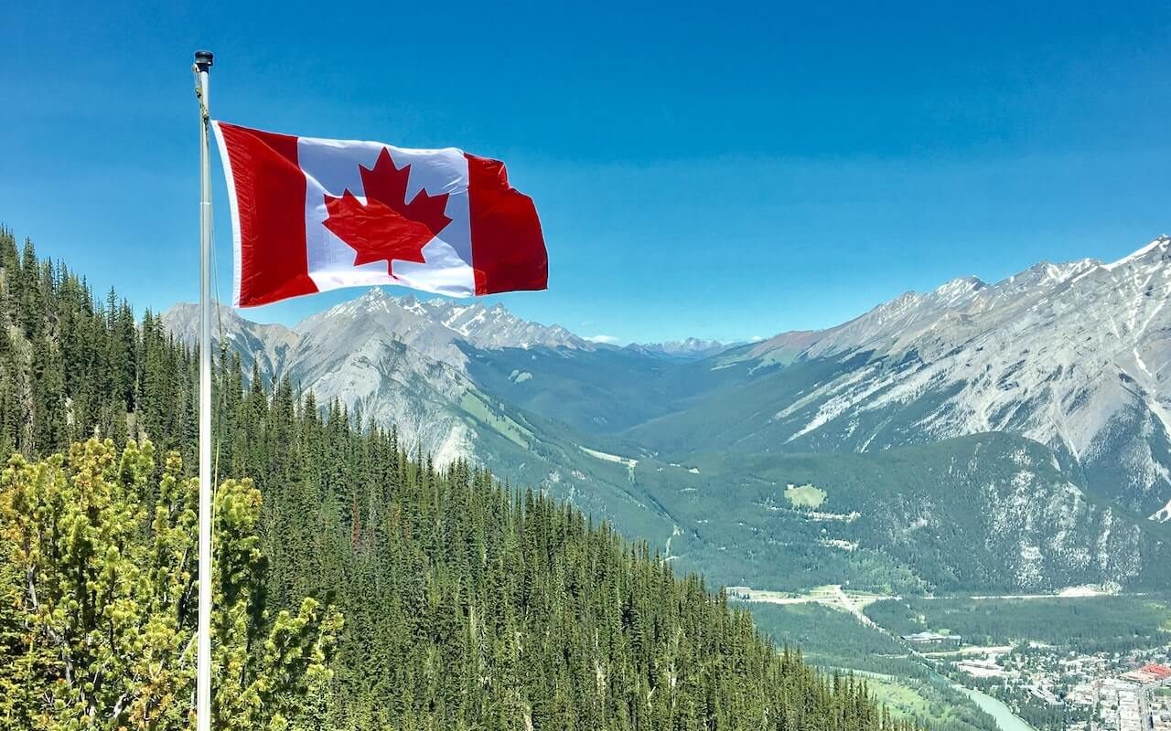 Image of the Canadian flag proudly waving over a picturesque landscape of trees and mountains.